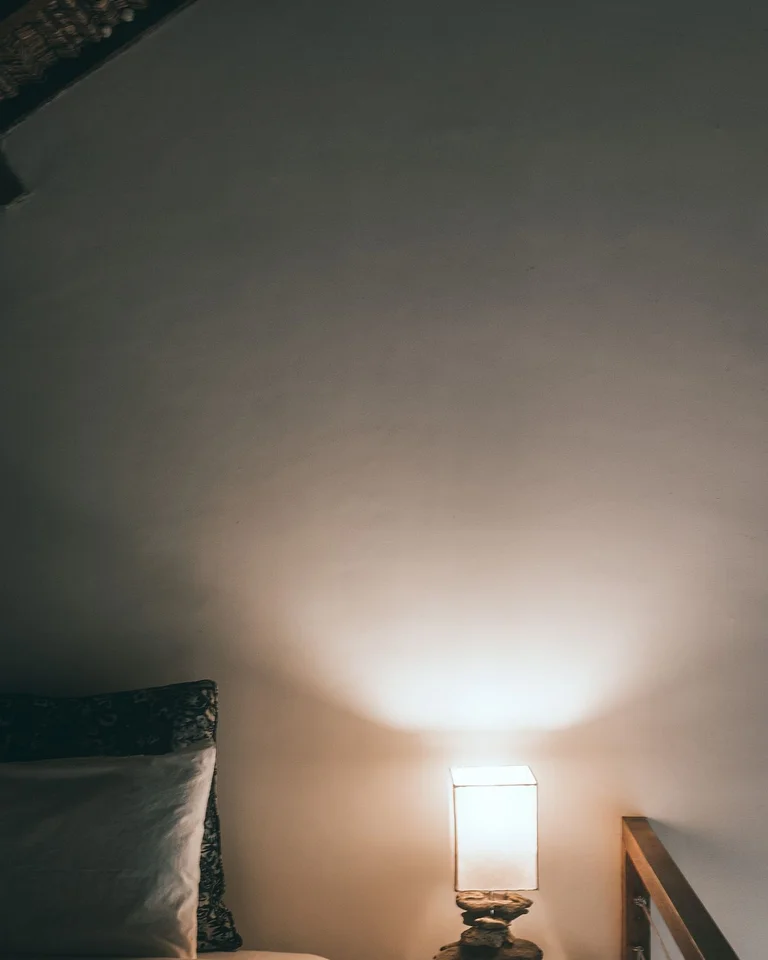 A warm bedside wall lamp glowing softly beside a bed in a dim bedroom