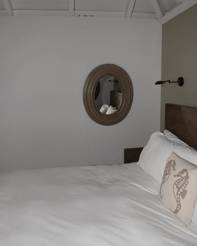 A cozy bedroom with a white bedspread and wooden headboard styled with a leaning mirror