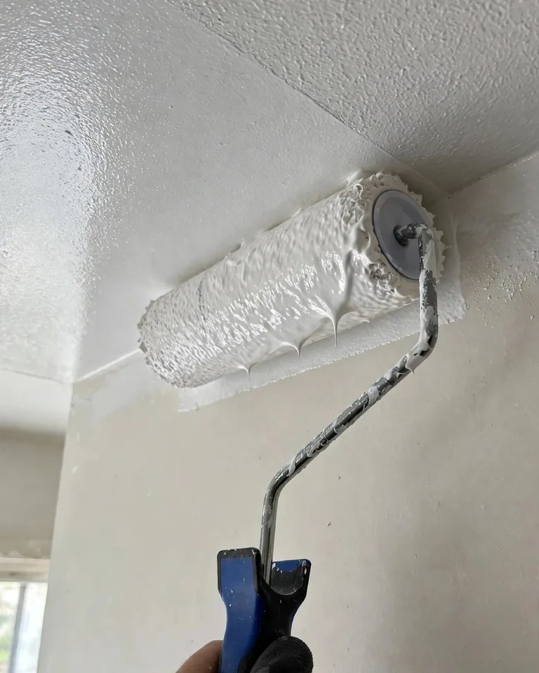 A paint roller applying flat white paint to a bedroom ceiling with a clean cut-in edge