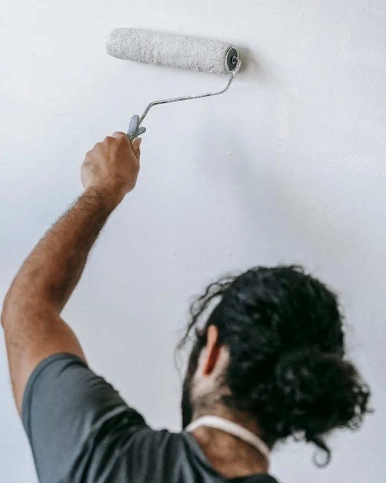 A person painting a bedroom wall white with a paint roller during a weekend makeover