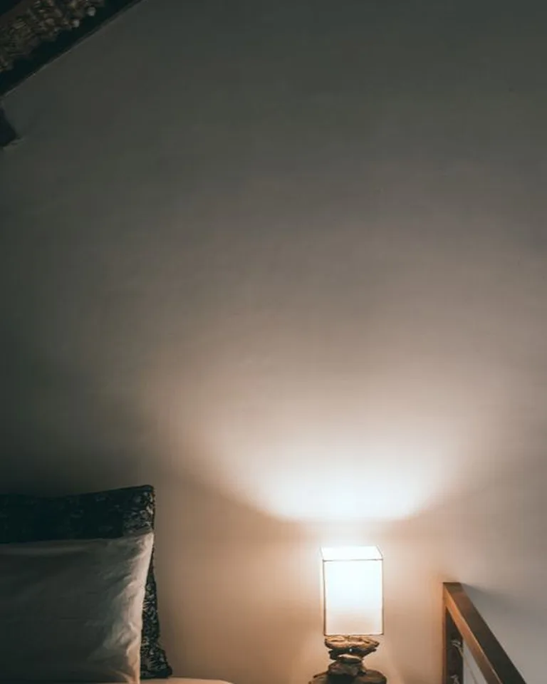 A warm bedside lamp glowing beside a bed in a cozy no-closet bedroom at dusk