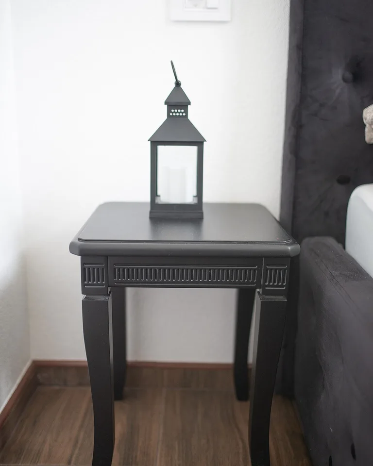 Black side table with a black lantern beside a bed with a dark headboard