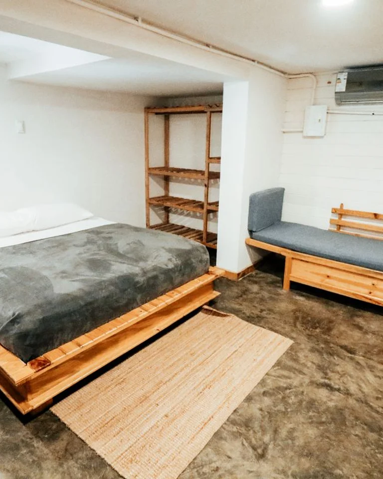 A small studio room with a wooden bed, a daybed seat, and a woven rug