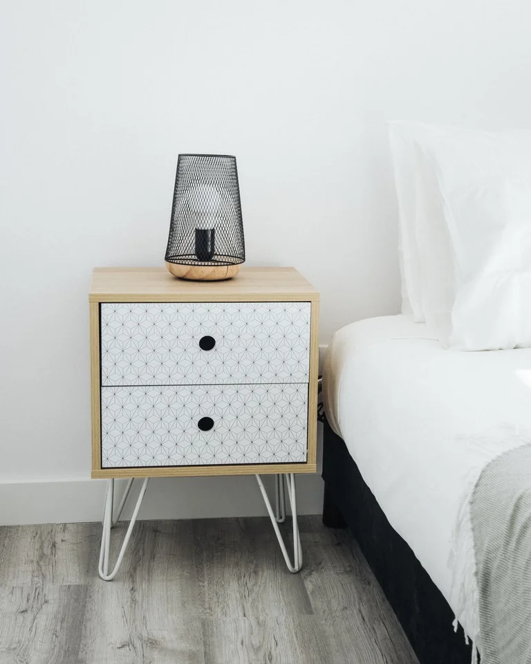 A light wood nightstand with white metal legs and a black mesh lamp beside a bed
