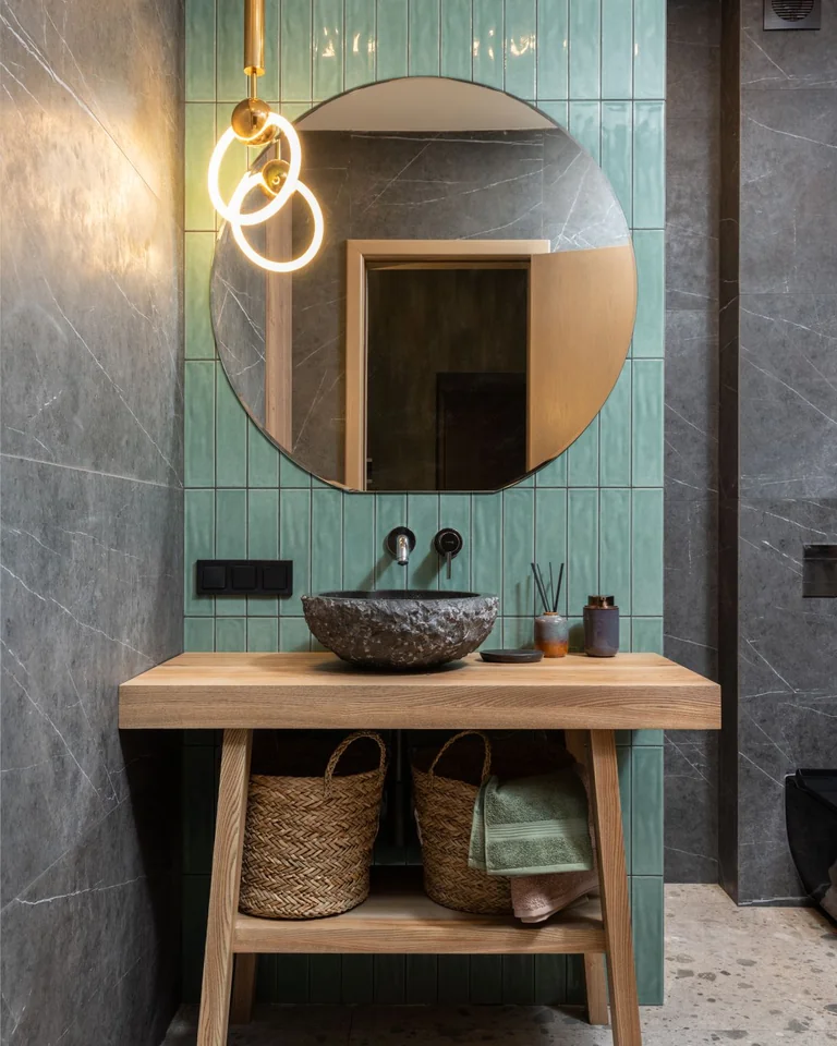 Bathroom with a green tiled accent wall behind a wooden vanity, stone vessel sink, and round mirror