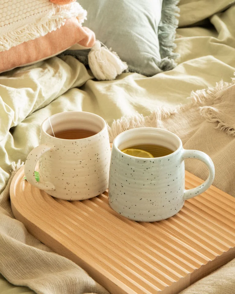 Two tea cups on a wooden tray on sage bedding with a terracotta-toned pillow